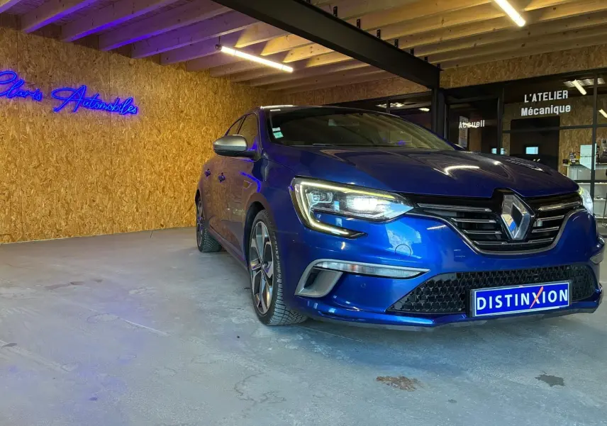 Renault Mégane 1.7 BlueDCI 150 GT Line bleu vue 3/4 avant droit, phare allumé en intérieur d'atelier.