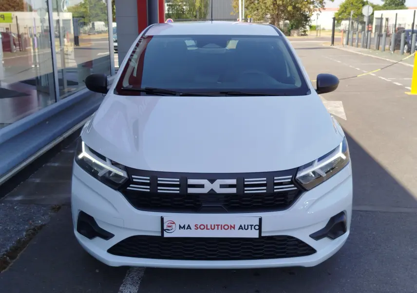 Vue avant d'une Dacia Sandero 2025 blanc glacier avec calandre noire et phares LED allumés, stationnée devant un bâtiment.