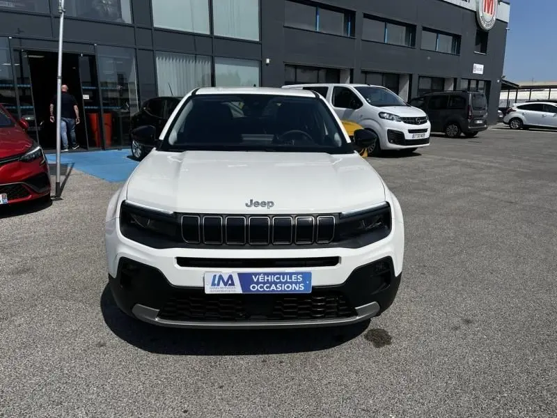 Vue avant d’un Jeep Avenger blanc électrique 2025 avec calandre noire et plaque « Véhicules Occasions » sur parking.