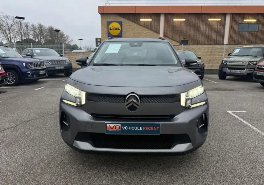 Vue frontale d'une Citroën C3 gris 2025 avec feux allumés et barres de toit visibles sur un parking.
