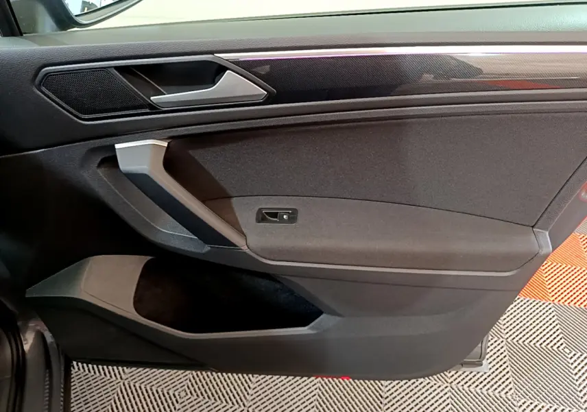 Vue rapprochée de la porte côté gauche intérieure du Volkswagen Tiguan gris clair, avec poignée argentée et garniture noire.