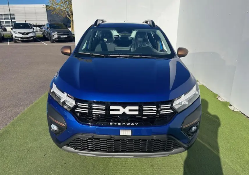 Vue avant d'une Dacia Sandero III Stepway bleu Iron avec calandre noire et détails brun cuivré sur un sol vert.
