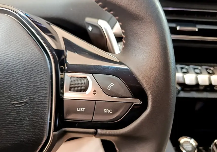Gros plan sur les commandes gauche du volant cuir noir du Peugeot 3008 gris foncé, avec tableau de bord flou en arrière-plan.