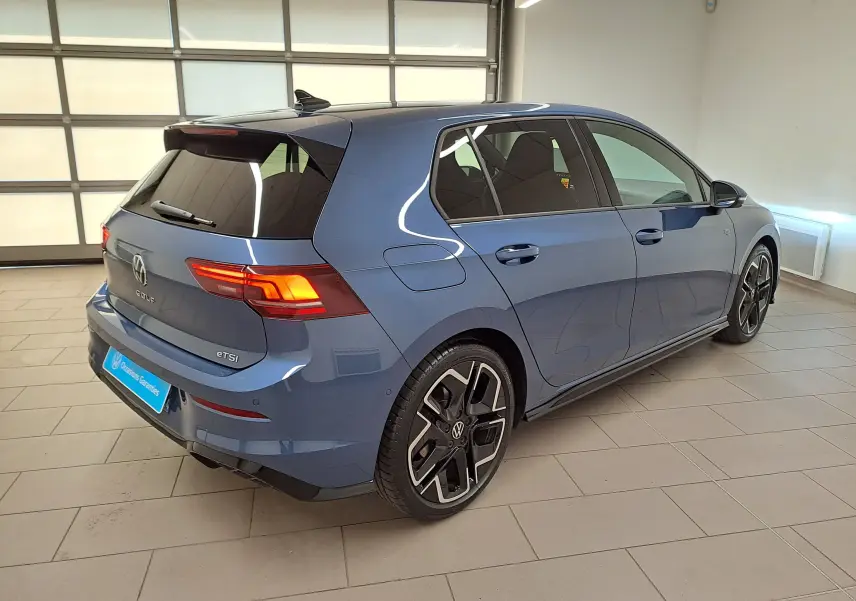 Vue 3/4 arrière droite d'une Volkswagen Golf 1.5 eTSI R-Line Bleu Anémone avec jantes noires et feux allumés.
