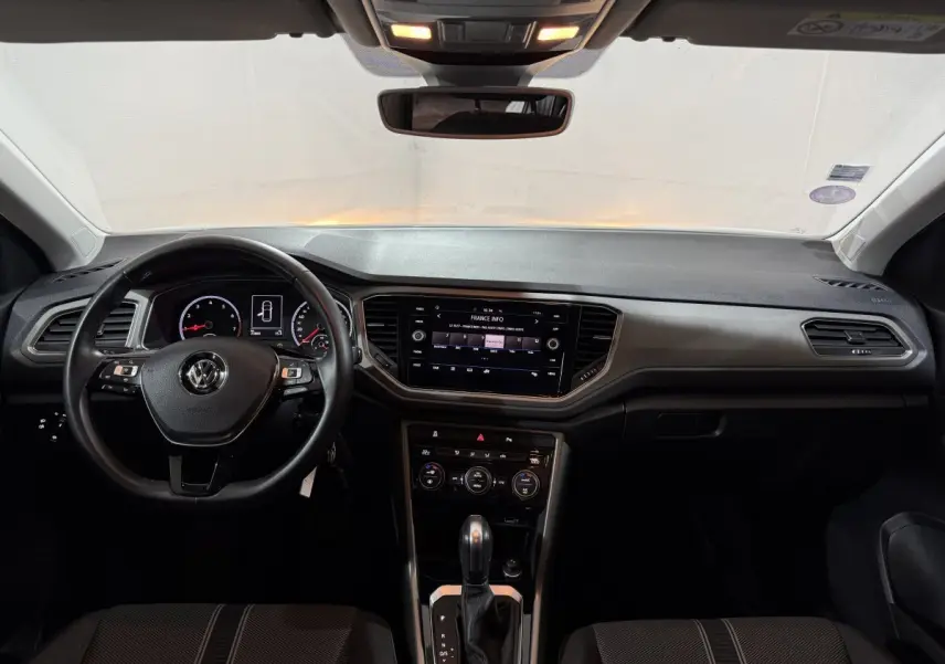 Vue intérieure frontale du tableau de bord et volant cuir noir du Volkswagen T-Roc blanc de 2020 avec écran tactile central allumé.