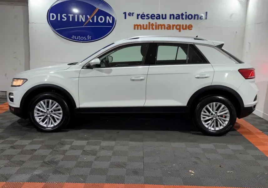 Profil côté gauche d'un Volkswagen T-Roc blanc 2020 avec jantes alliage et barres de toit noires en intérieur showroom.