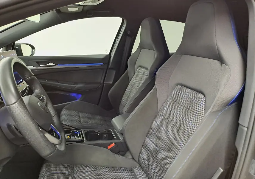 Vue intérieure côté conducteur de la Volkswagen Golf 1.5 eHybrid 2025 avec sièges sport gris à motif tartan et éclairage d'ambiance bleu.