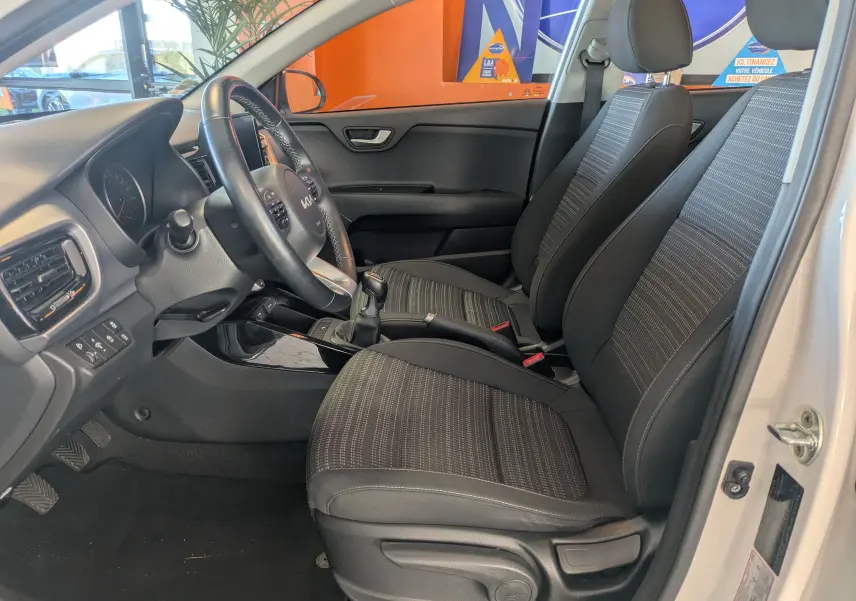 Vue intérieure côté conducteur du Kia Stonic blanc 2022, montrant les sièges tissu noirs et la boîte manuelle.