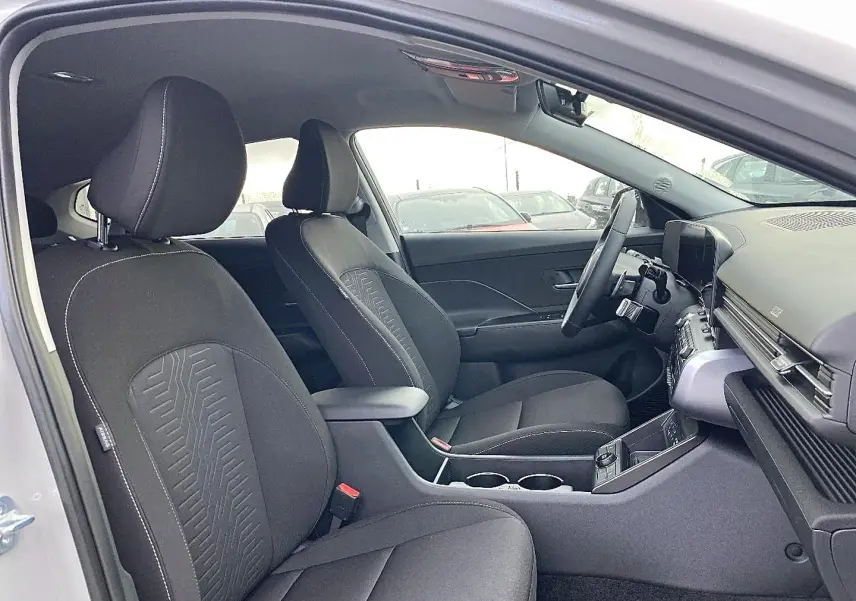 Intérieur de la Hyundai Kona 2025 vue côté conducteur, sièges tissu noirs avec accoudoir central et tableau de bord moderne.