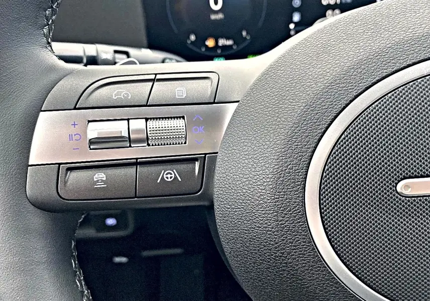 Gros plan sur les commandes à gauche du volant cuir du Hyundai Kona gris, avec tableau de bord numérique en arrière-plan.
