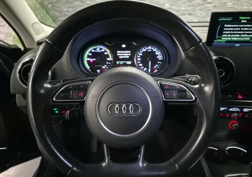 Vue intérieure centrée sur le volant cuir noir de l'Audi A3 Sportback avec tableau de bord et écran multimédia allumé.