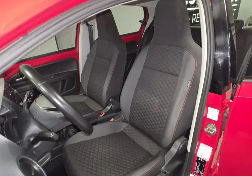 Vue intérieure côté conducteur d'une Volkswagen Up rouge, sièges avant en tissu noir avec surpiqûres rouges.