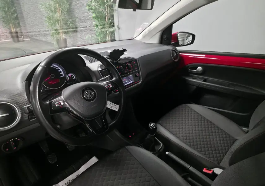 Intérieur avant droit d'une Volkswagen Up rouge, volant cuir multifonctions et sellerie tissu avec surpiqûres rouges.