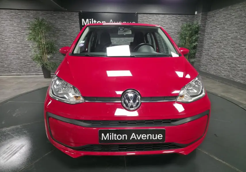 Vue frontale d'une Volkswagen Up rouge vif avec logo VW centré et plaque "Milton Avenue" en intérieur showroom.