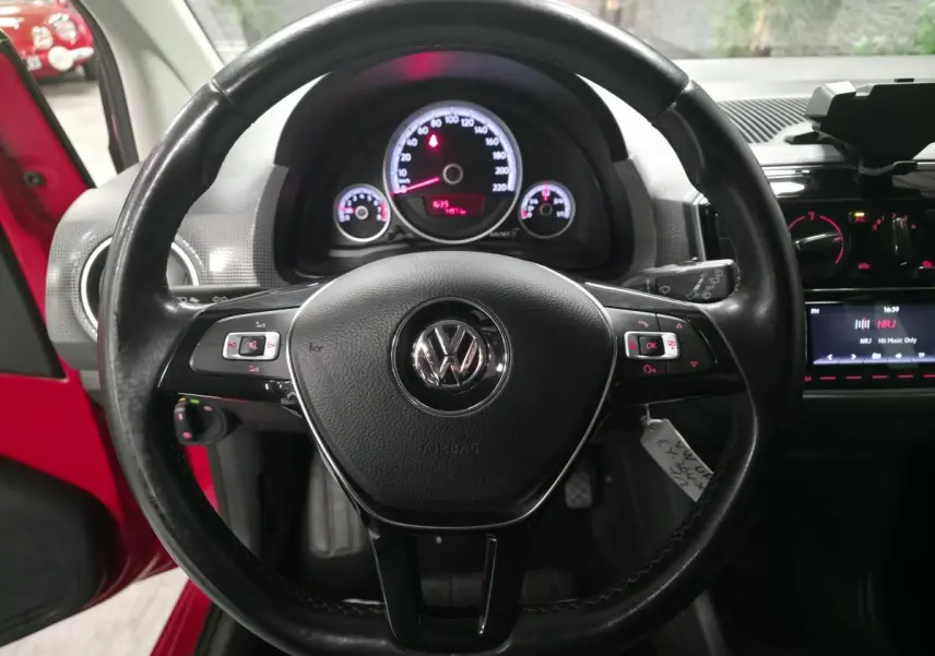 Vue intérieure centrée sur le volant cuir multifonctions d'une Volkswagen Up rouge, avec tableau de bord éclairé.