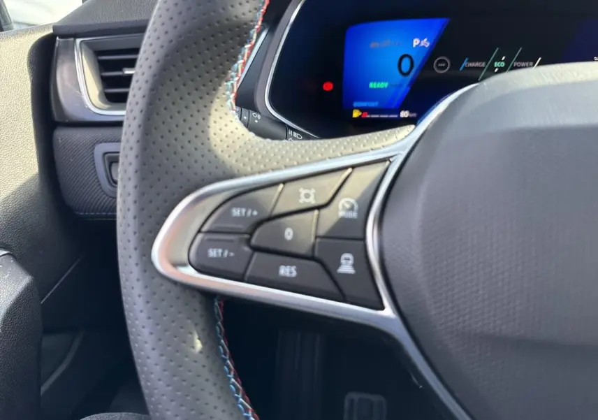 Gros plan sur le volant perforé côté gauche avec commandes multifonctions et tableau de bord numérique bleu du Renault Symbioz blanc nacré.