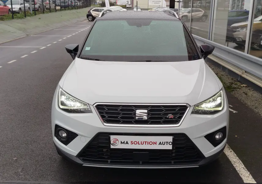 Vue frontale d'un SEAT Arona blanc avec toit gris magnétique, phares allumés et calandre noire hexagonale.