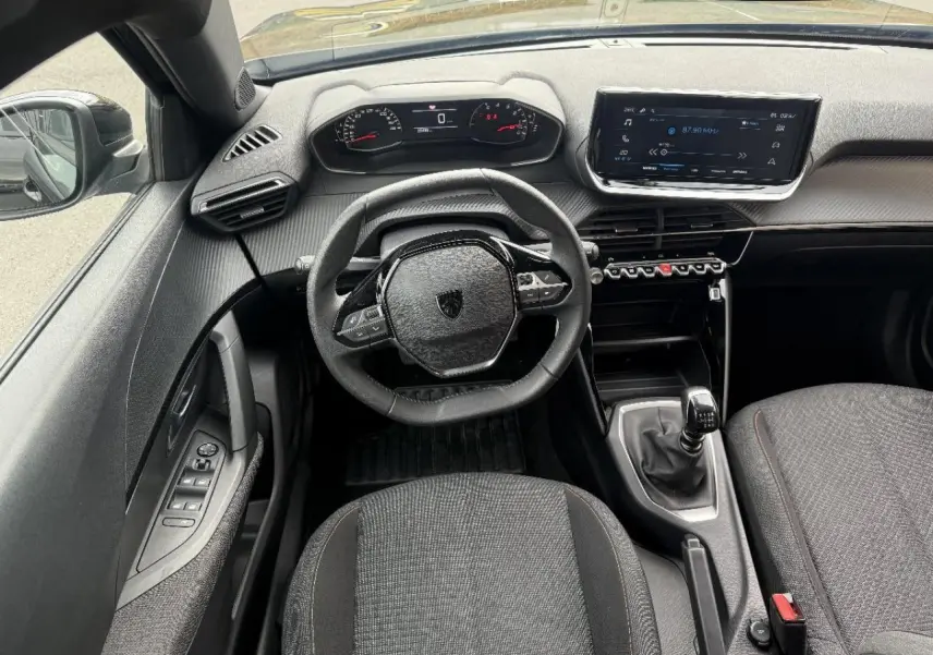 Vue intérieure de face du poste de conduite d'une Peugeot 2008 noire, avec volant compact, tableau de bord numérique et boîte manuelle.