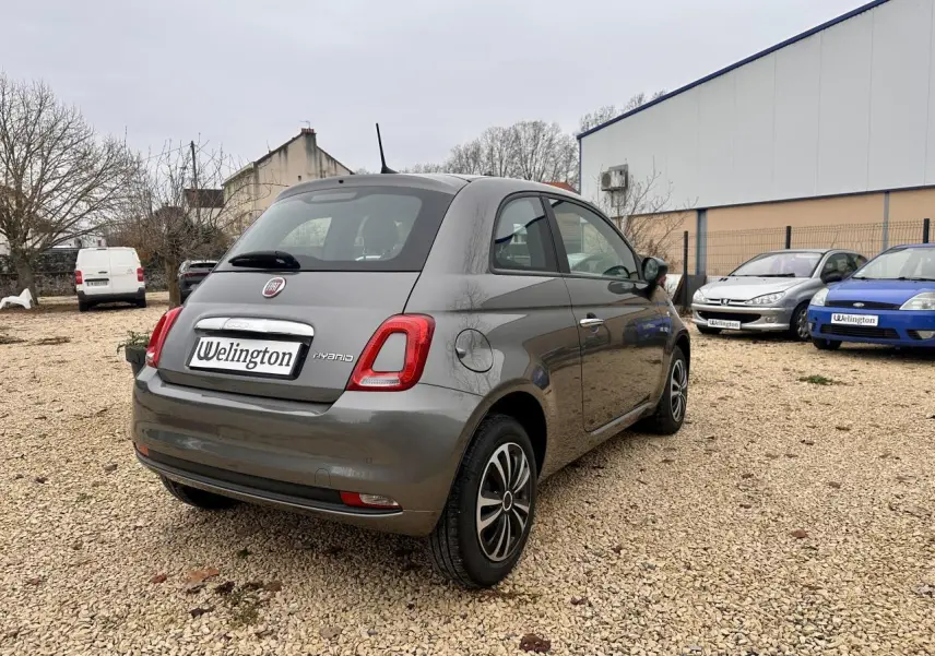 Fiat 500 gris foncé vue 3/4 arrière droit sur un sol gravillonné avec plusieurs voitures en arrière-plan.