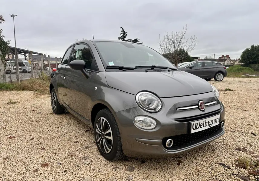 Fiat 500 gris foncé vue 3/4 avant droit sur un sol gravillonné avec jantes alliage et phares ronds.