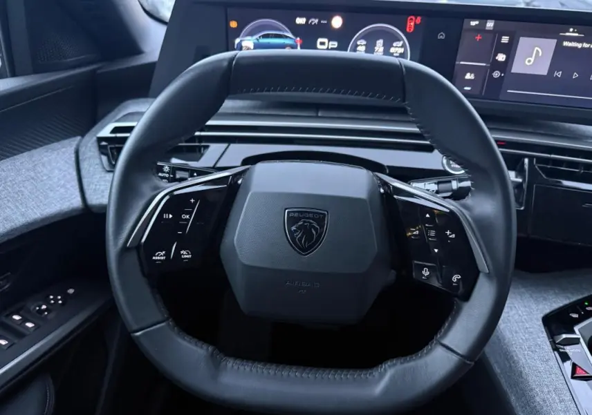 Vue rapprochée du volant cuir noir du Peugeot 3008 Hybrid 145 Allure 2025 avec cockpit numérique en arrière-plan.