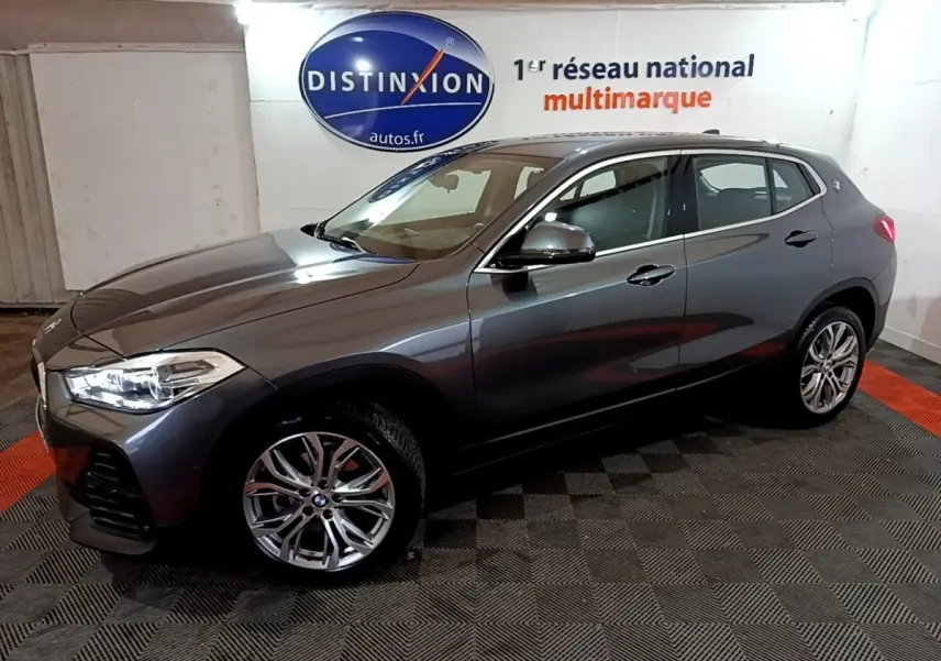 BMW X2 sDrive18i gris foncé vue de profil côté gauche, avec jantes alliage et phares allumés dans un showroom.