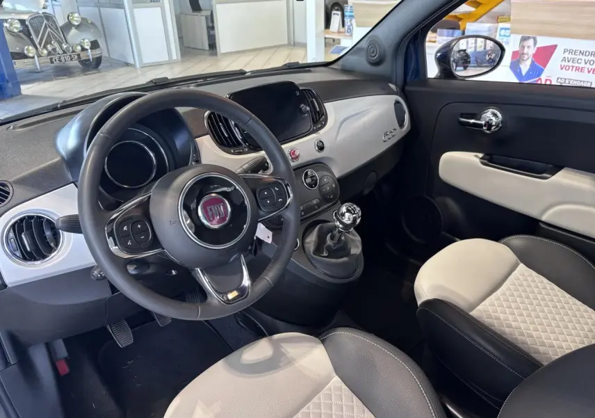 Vue intérieure côté conducteur de la Fiat 500 avec volant simili-cuir, tableau de bord blanc et sièges bicolores matelassés.