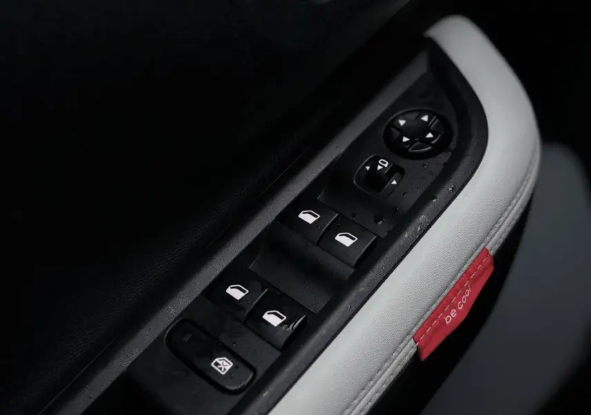 Gros plan sur les commandes des vitres électriques côté conducteur avec surpiqûres blanches et étiquette rouge "be cool" dans l'habitacle.