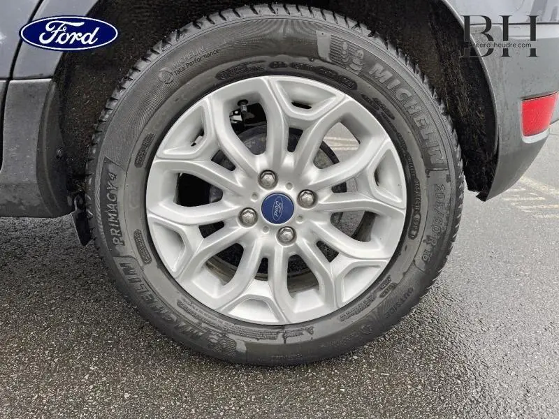 Gros plan sur la roue avant droite avec jante alu argentée et logo Ford, partie arrière Smoke visible sur sol humide.