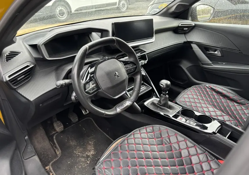 Vue intérieure côté conducteur de la Peugeot 208 GT 2021 jaune, avec volant compact, boîte manuelle et sièges noirs matelassés rouges.