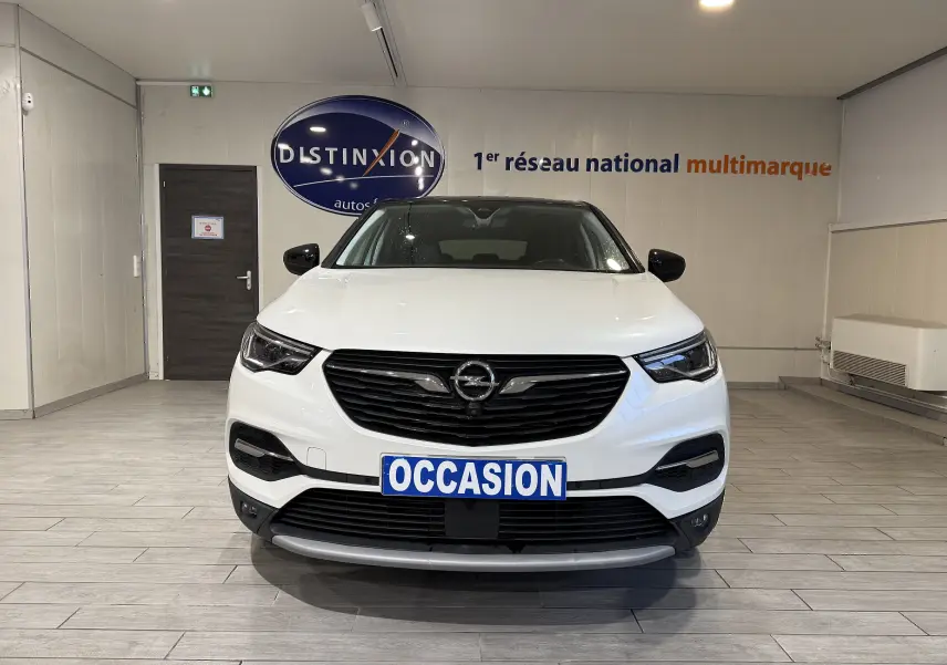 Vue frontale d’un Opel Grandland X blanc hybride rechargeable, avec calandre noire et plaque "OCCASION" en intérieur showroom.