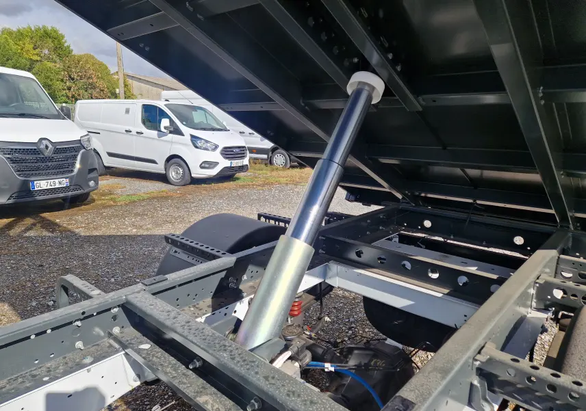 Vue rapprochée sous la benne relevée d’un Ford Transit blanc, mettant en valeur le vérin hydraulique et le châssis acier.