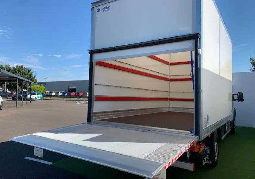 Vue arrière d'un FIAT Ducato blanc avec hayon élévateur déployé et caisse fourgon vide ouverte sur un parking.