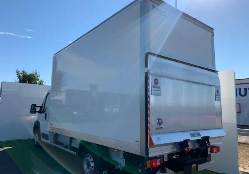 Fiat Ducato Fourgon blanc vu de l'arrière droit, avec hayon élévateur et caisse fermée sous un ciel bleu.