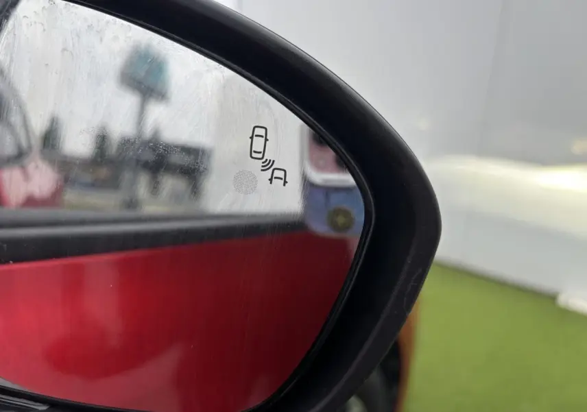 Gros plan sur le rétroviseur extérieur gauche d'une Renault Clio rouge flamme, avec icône d'alerte d'angle mort visible.