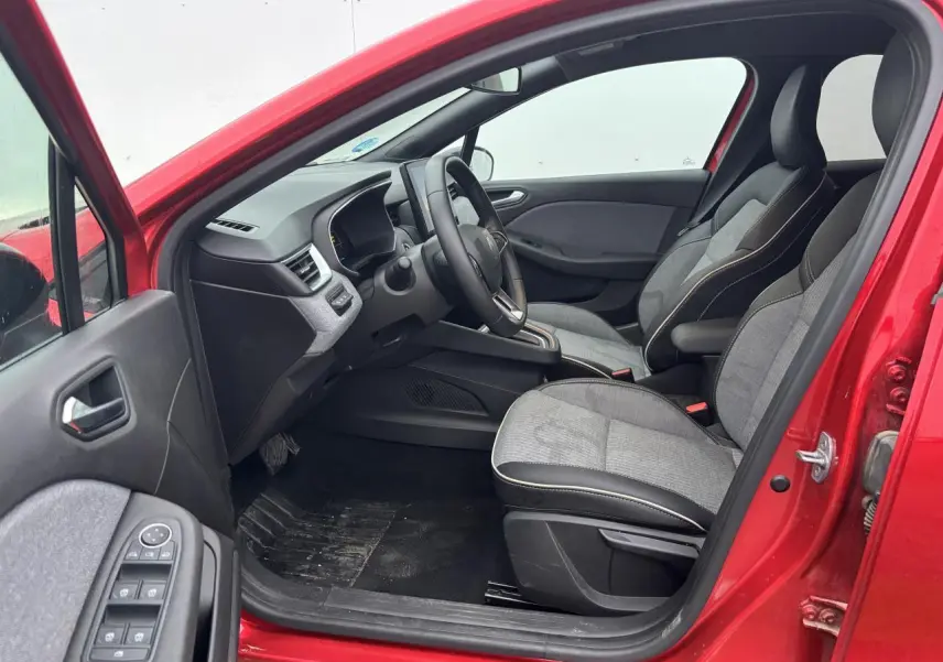 Vue intérieure côté conducteur de la Renault Clio V rouge flamme, avec sièges gris et tableau de bord moderne.