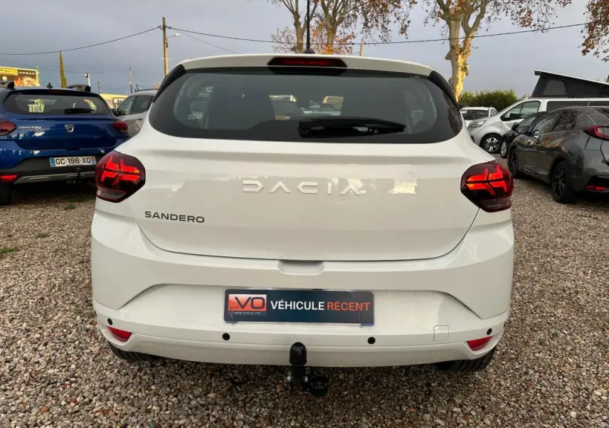 Vue arrière d'une Dacia Sandero blanche 2024 avec feux arrière allumés et attelage de remorque visible.