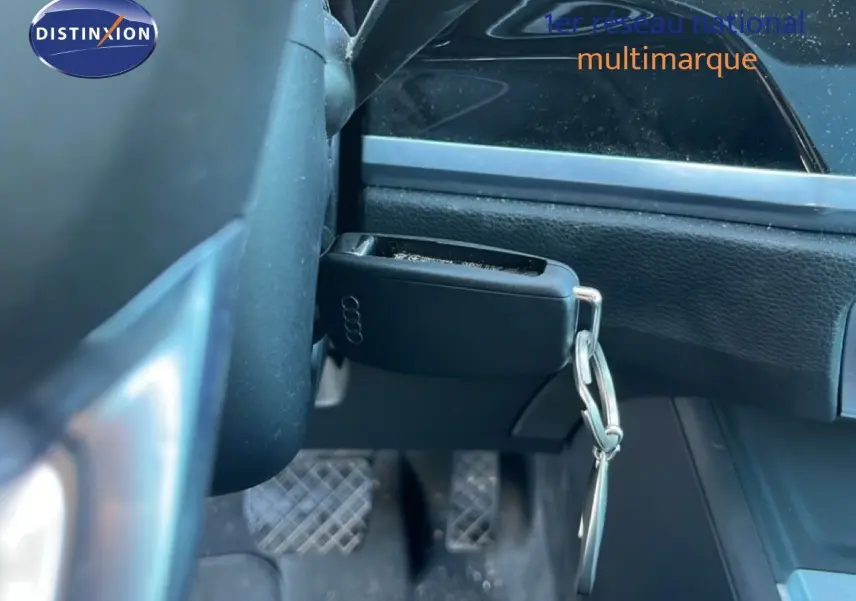 Gros plan sur la clé noire Audi suspendue près du volant, avec les pédales et la console noire en arrière-plan.