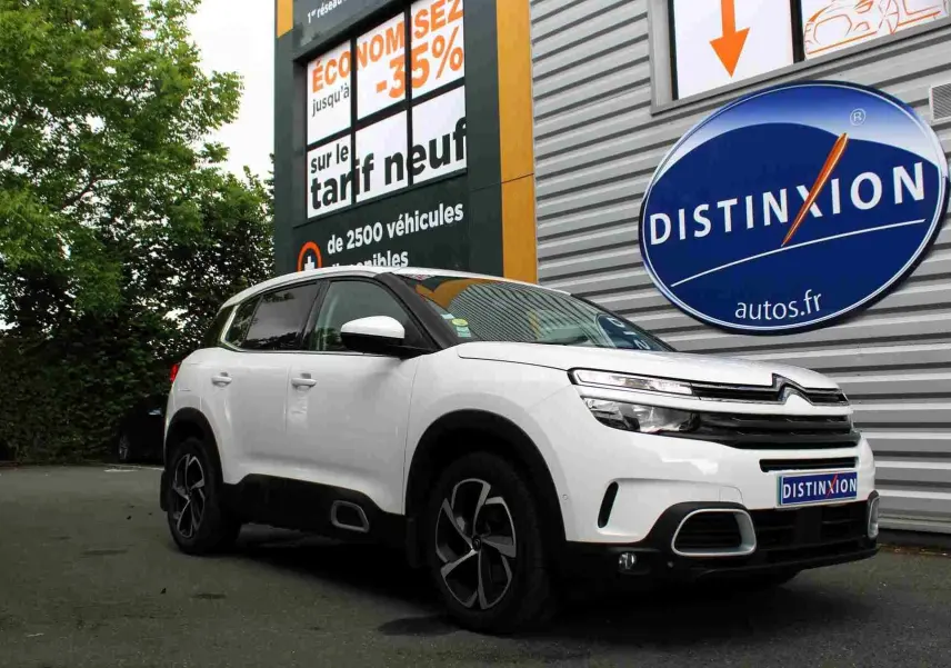 Vue 3/4 avant d'un Citroën C5 Aircross blanc avec jantes noires et toit noir, stationné devant un panneau publicitaire.