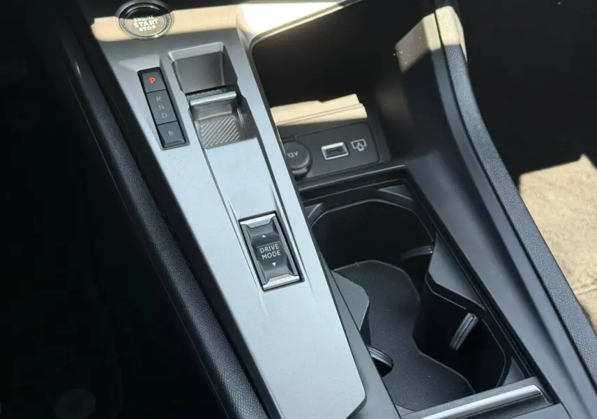Vue en plongée sur la console centrale grise de la Peugeot e-308 2025 avec sélecteur de mode de conduite et prises USB/12V.