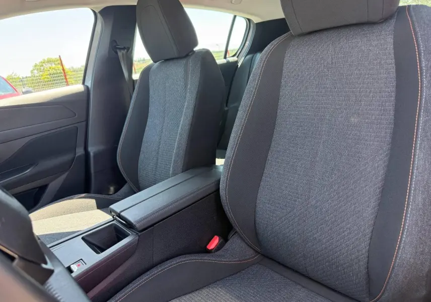 Gros plan sur les sièges avant gris tissu avec surpiqûres orange et accoudoir central de la Peugeot e-308 2025.