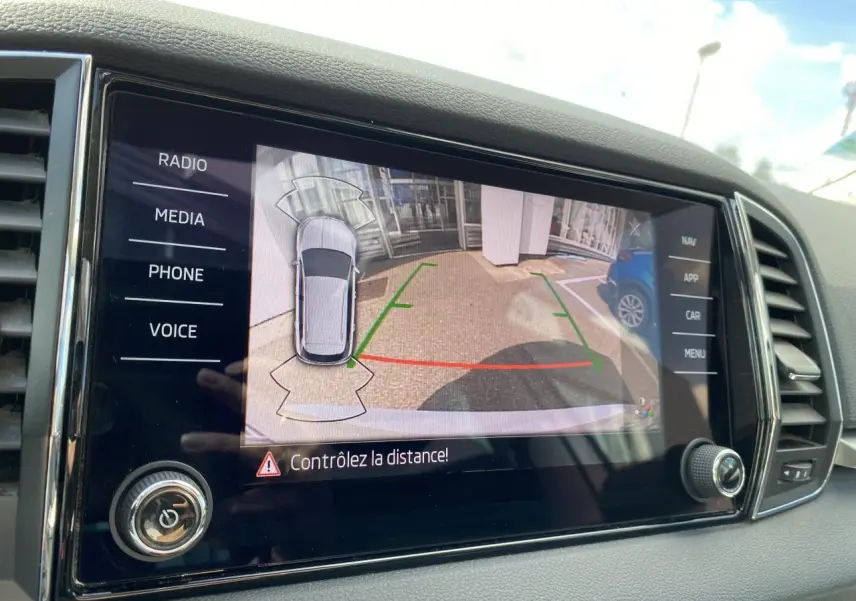 Écran tactile intérieur montrant la caméra de recul avec vue aérienne du Skoda Karoq blanc Moon métallisé en stationnement.