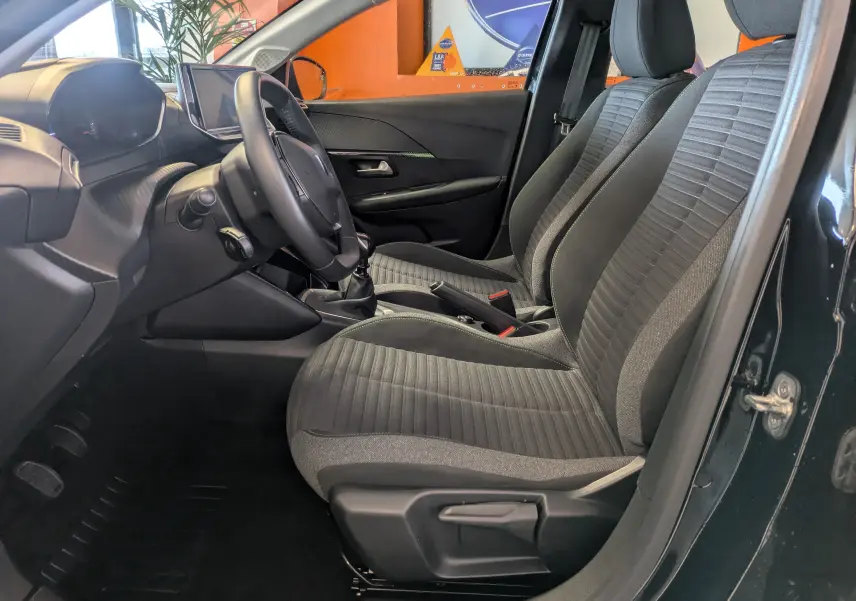 Vue intérieure côté conducteur de la Peugeot 208 noire, montrant les sièges tissu gris et le volant cuir avec boîte manuelle.