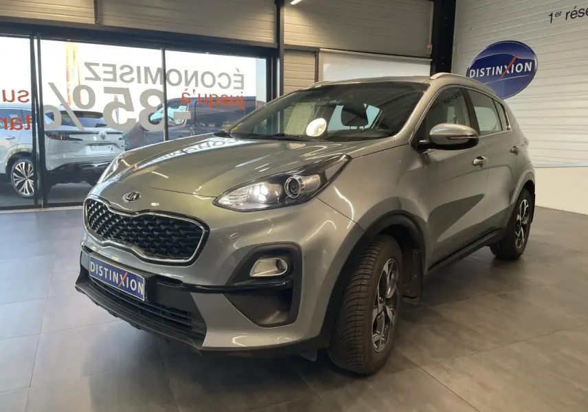 KIA Sportage gris clair en 3/4 avant droit, avec calandre noire mat et jantes alliage 16 pouces visibles.