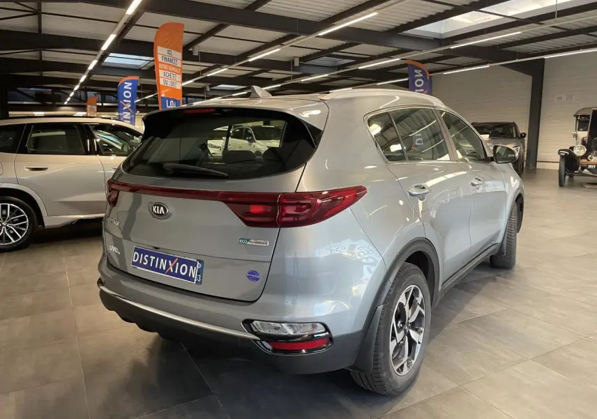 Vue 3/4 arrière droite du KIA Sportage gris clair métallisé en showroom avec jantes alliage et becquet arrière intégré.