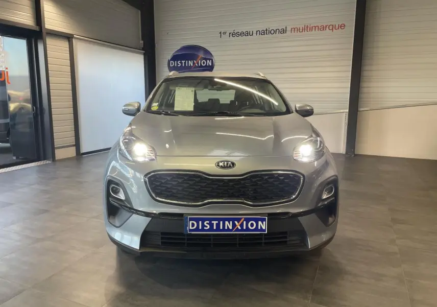 Vue avant d'un KIA Sportage gris clair avec phares allumés, calandre noire et plaque Distinxion en intérieur showroom.