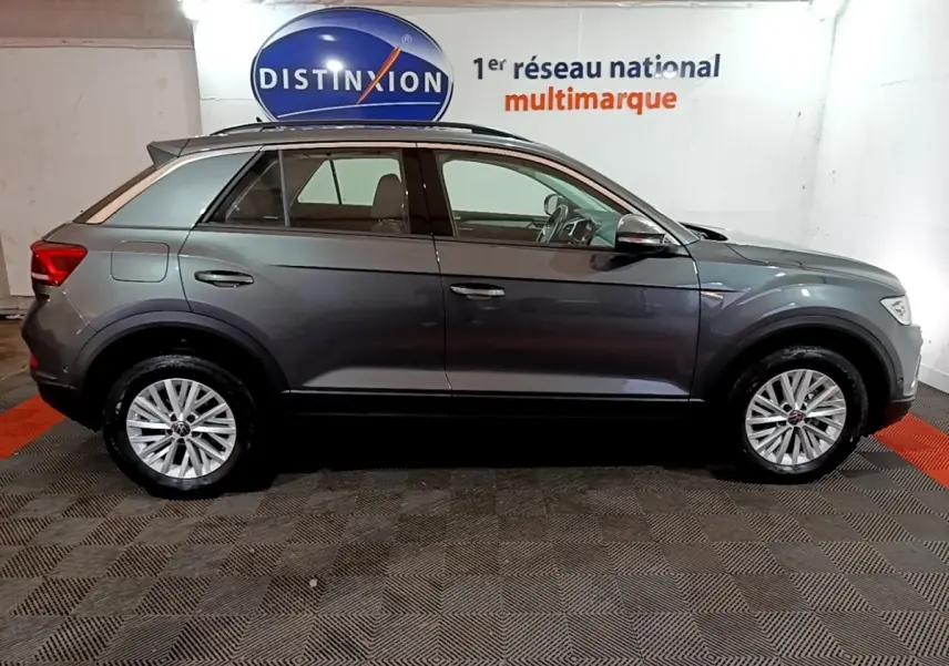 Vue de profil côté gauche d'un Volkswagen T-Roc gris foncé 2022 en intérieur avec jantes alliage et barres de toit noires.
