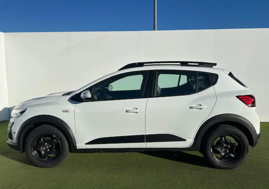 Vue latérale droite d'une Dacia Sandero Stepway blanche Arctique avec barres de toit noires et jantes foncées.