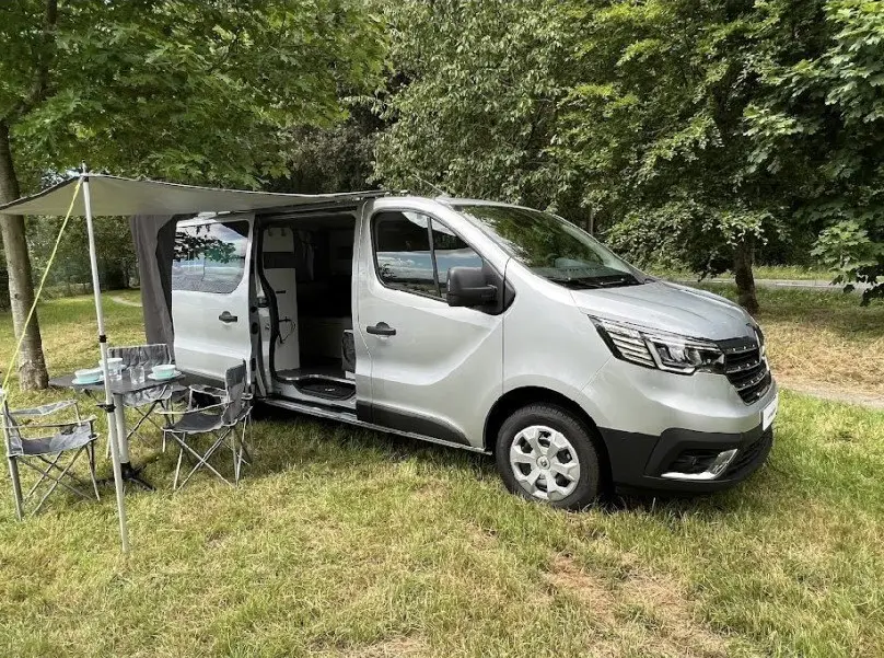 Renault Trafic Fourgon gris en 3/4 avant droit avec porte latérale ouverte et auvent déployé en pleine nature.