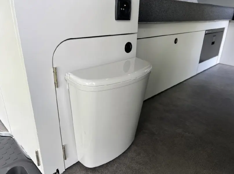 Détail intérieur du fourgon Renault Trafic aménagé, montrant une poubelle blanche intégrée près d’un meuble gris clair.